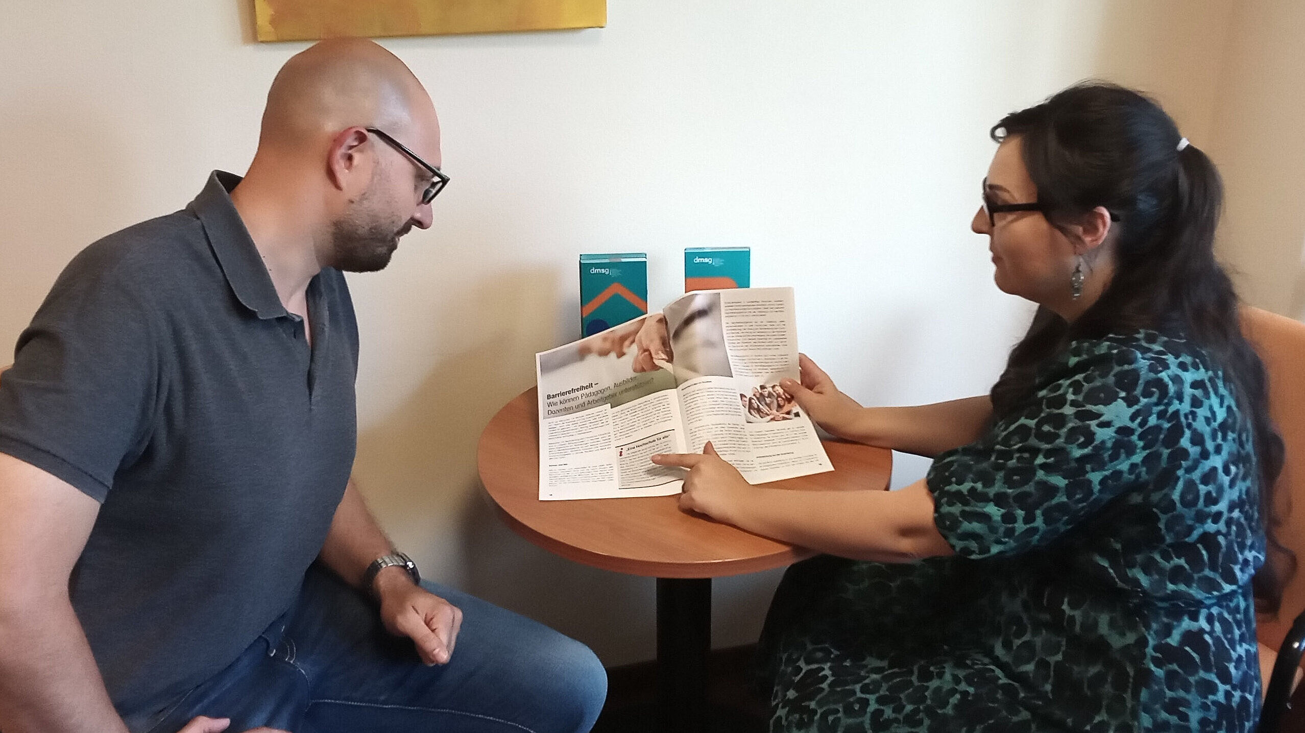 Ein Mann und eine Frau an einem Tisch. Die Frau zeigt dem Mann etwas in einer Broschüre.
